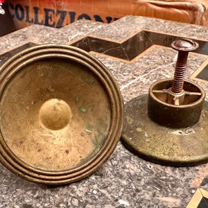 May include: Two antique brass objects. One is a circular base with concentric rings and a central knob. The other has a similar base with a threaded rod and a screw head. The objects have a weathered, aged appearance.