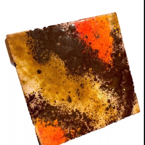 May include: A square ceramic tile with a brown, orange, and yellow abstract design. The tile has a speckled, textured surface.