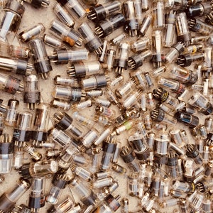 May include: A large collection of vintage vacuum tubes, also known as valves, in various sizes and shapes. The tubes are made of glass and have metal pins at the bottom. They are arranged in a random pattern on a light brown surface.