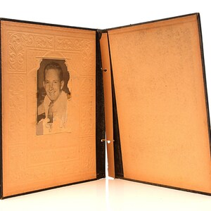May include: An open black photo album with a black and white photo of a man in a suit and tie. The album has a decorative embossed design on the front cover.