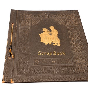 May include: A vintage black scrapbook with a gold embossed design and the words "Scrap Book" on the front cover. The cover features a gold silhouette of two people.
