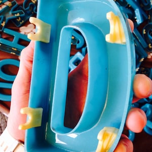 Peut inclure: Une grande lettre D en plastique bleu vif avec des languettes en plastique jaunes sur les c&ocirc;t&eacute;s. La lettre est tenue par une main.
