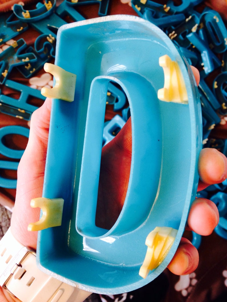 Peut inclure: Une grande lettre D en plastique bleu vif avec des languettes en plastique jaunes sur les c&ocirc;t&eacute;s. La lettre est tenue par une main.