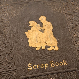 May include: A vintage brown leather scrapbook with gold embossed details and a gold inscription that reads "Scrap Book". The cover features a gold silhouette of a couple.