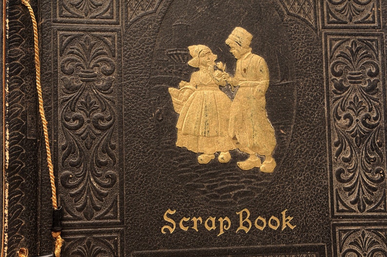 May include: A vintage leather-bound scrapbook with a gold embossed design featuring two figures in traditional Dutch clothing. The title "Scrap Book" is written in gold lettering on the front cover.