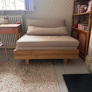 May include: A light beige linen futon couch with a wooden frame and legs. The couch is in a room with a patterned wallpaper and a rug with a multi-colored pattern.