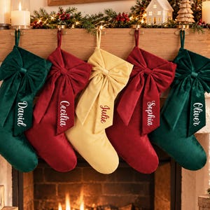 May include: Five velvet Christmas stockings hanging in a row, featuring large bows and personalised names embroidered on the streamers. The stockings are coloured dark green, deep red, and gold, with the names David, Cecilia, Julie, Sophia, and Oliver.