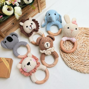May include: A selection of handmade crochet baby rattles in animal shapes, such as a lion, elephant, koala, bear, and bunny. Each rattle has a wooden ring and is made in soft, neutral colours. These rattles are designed for infants and toddlers.