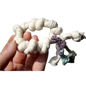May include: A handmade, off-white fabric dog toy in a circular shape, with a knotted design. Accented with purple, teal, and gray knotted fabric. The toy is held in a person's hand.