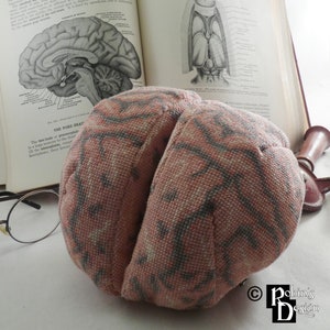 May include: A pink and gray fabric brain model with a detailed anatomical design. The brain is sitting on a table with a book and glasses.