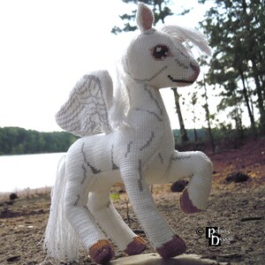 Puede incluir: Un caballo alado blanco de crochet con pezuñas marrones, de pie sobre una roca en un entorno de bosque. El caballo tiene una crin y una cola blancas. Las alas están detalladas con puntadas grises. El caballo está mirando hacia la derecha. El texto "Robin's Design" es visible en la esquina inferior derecha de la imagen.