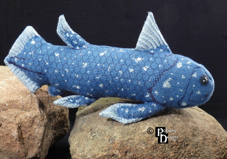 May include: A blue and white crocheted stuffed animal resembling a prehistoric fish with a spotted pattern. The fish is posed on two brown rocks.