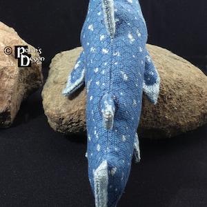 May include: A blue and white knitted whale with a spotted pattern. The whale is lying on a rock.
