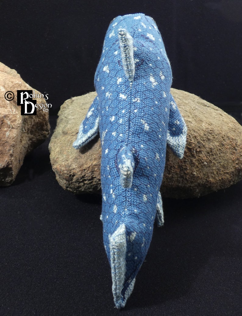 May include: A blue and white knitted whale with a spotted pattern. The whale is lying on a rock.