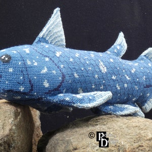 May include: A blue and white crocheted stuffed fish with a spotted pattern. The fish is posed on two gray rocks.