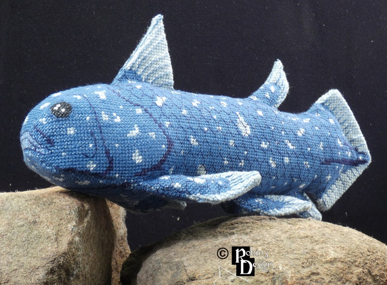 May include: A blue and white crocheted stuffed fish with a spotted pattern. The fish is posed on two gray rocks.