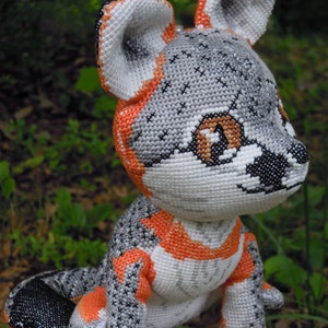 May include: A grey and orange crocheted stuffed animal resembling a fox with large ears and brown eyes. The animal is sitting on a rock with a green background.