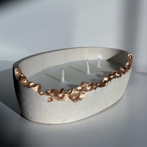 Puede incluir: Una vela blanca de forma ovalada en un recipiente de hormigón con tres mechas. El borde superior del recipiente presenta un acabado dorado metálico texturizado que imita la apariencia de un borde rugoso y dentado, contrastando con la cera blanca.