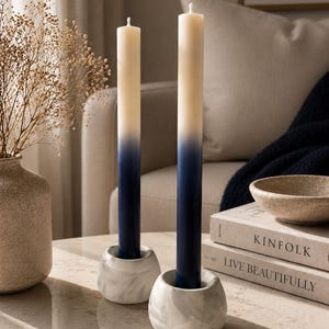 May include: Two ombre candles in white marble holders. The candles transition from cream to navy blue. The holders have a textured, marbled appearance. The candles are displayed on a light-coloured surface.