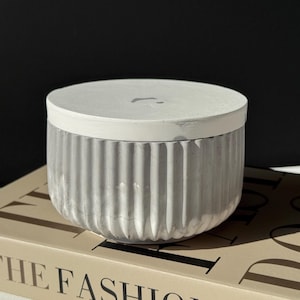 May include: A gray, ribbed concrete container with a white lid. The container is round and sits on a book with the words "THE FASHION BOOK" printed on the cover. The background is black.