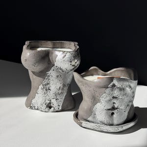 May include: Two gray concrete torso-shaped candle holders with silver leaf accents. The candles are filled with white wax. One candle holder is on a matching saucer. The background is black and white.