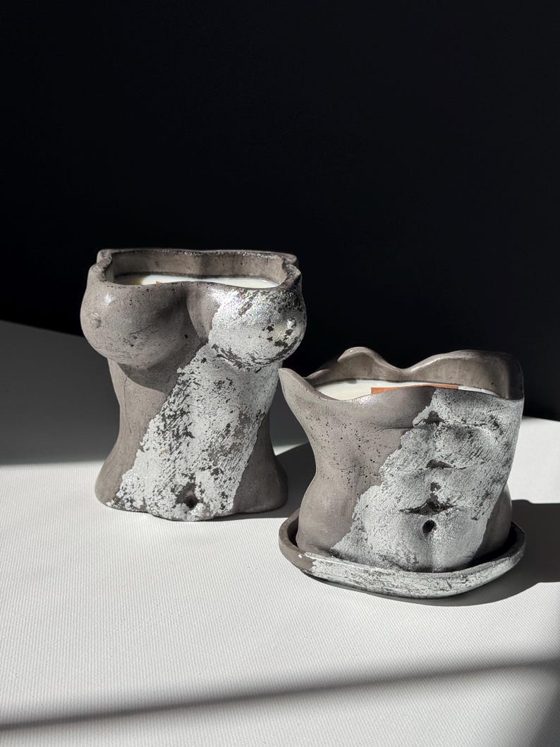 May include: Two gray concrete torso-shaped candle holders with silver leaf accents. The candles are filled with white wax. One candle holder is on a matching saucer. The background is black and white.