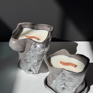 May include: Two gray concrete candles shaped like torsos. The candles have a silver metallic finish on the surface and are filled with white wax. Each candle has a wooden wick. The candles are on a white surface.