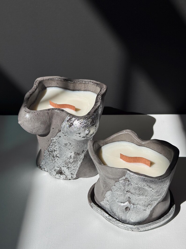May include: Two gray concrete candles shaped like torsos. The candles have a silver metallic finish on the surface and are filled with white wax. Each candle has a wooden wick. The candles are on a white surface.