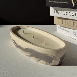 May include: Oval-shaped candle with three wicks, set in a marbled gray and cream-colored concrete container. The candle is a light cream color and sits on a white surface. Books are stacked in the background.