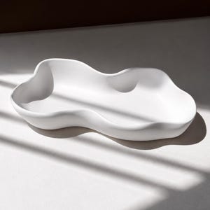 Może przedstawiać: Matowa biała taca ceramiczna o nieregularnej, organicznej i falistej sylwetce. Naczynie posiada gładką, ciągłą powierzchnię z zakrzywionymi, podniesionymi krawędziami, które tworzą płynny, chmurowy kształt, zapewniając płytkie wgłębienie na drobne przedmioty.