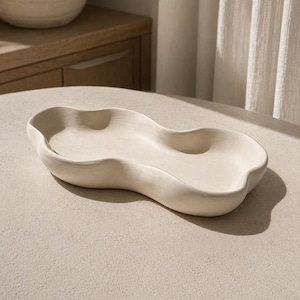 May include: A cream-coloured, organically shaped ceramic tray with a wavy design. The tray has a smooth surface and is set on a neutral-toned surface. The design features a series of gentle curves and indentations, creating a unique aesthetic.