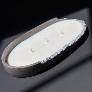 Puede incluir: Una vela ovalada de tres mechas en un recipiente de hormigón gris oscuro. El borde exterior del recipiente presenta un acento metálico texturizado de color plateado en un lado, que contrasta con la superficie lisa de cera color blanco roto.