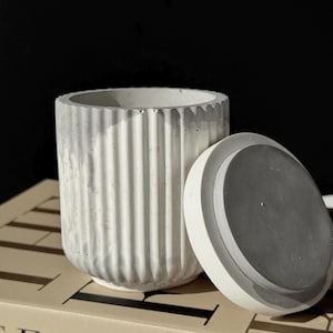 May include: A cylindrical, ribbed concrete container with a matching lid. The container is light gray with vertical ridges and a white rim. The lid is off-white with a gray interior. The container is sitting on a book with the word "FASHION" visible.