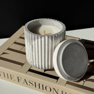 Peut inclure: Un bougeoir en béton gris avec un extérieur strié et un couvercle assorti. La bougie à l'intérieur est blanche avec une mèche en bois. Le bougeoir est posé sur un livre avec les mots "THE FASHION BOOK" imprimés sur la couverture.