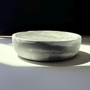 May include: A round, grey concrete bowl with a smooth, marbled appearance. The bowl has a low profile and a wide opening. The colour is a mix of light and dark grey, with a subtle, natural texture. The bowl is sitting on a white surface.