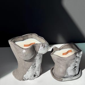 May include: Two gray concrete candles, one shaped like a torso and the other a smaller, rounded shape. Both have a white wax fill and a wooden wick. The torso candle has silver accents. The candles sit on a white surface.