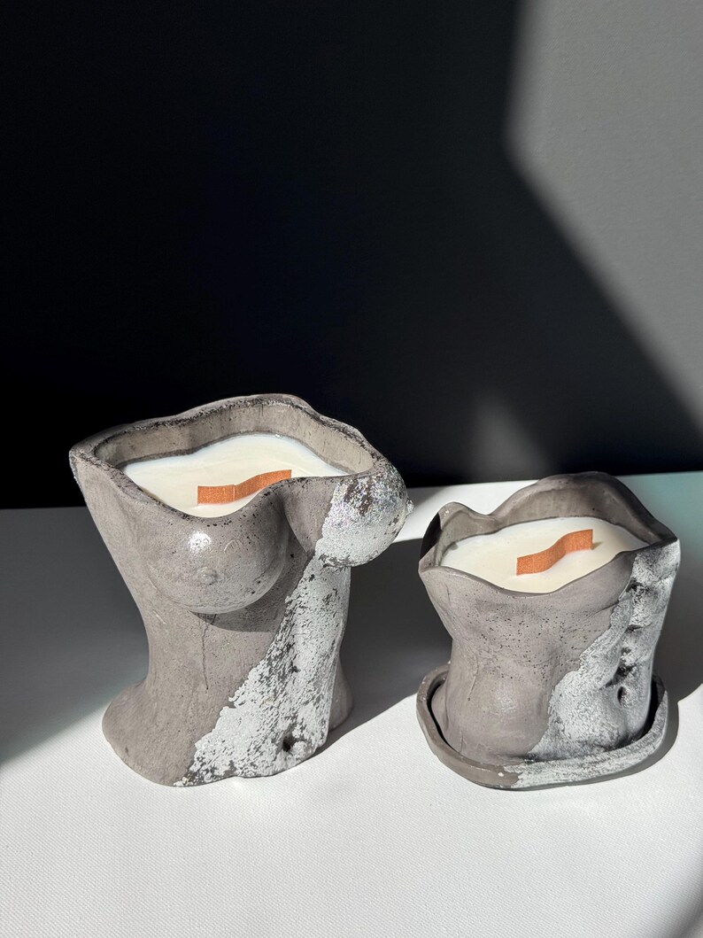 May include: Two gray concrete candles, one shaped like a torso and the other a smaller, rounded shape. Both have a white wax fill and a wooden wick. The torso candle has silver accents. The candles sit on a white surface.