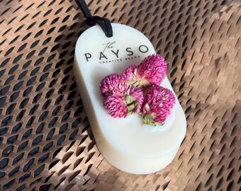 Puede incluir: Un ambientador ovalado blanquecino con el texto "The PAYSO CREATIVE BRAND" y tres flores rosas secas. Se adjunta un cordón negro para colgar. El ambientador está sobre una superficie marrón texturizada.