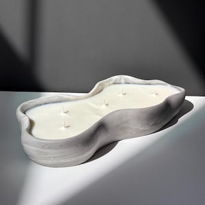 May include: A large, grey, concrete candle holder with a wavy, organic shape. The holder is filled with white wax and has four wicks.