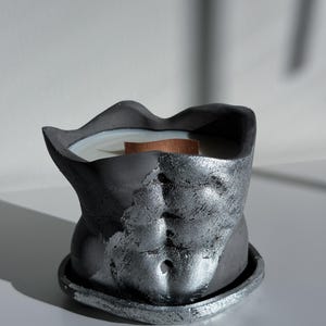 May include: A dark gray candle in the shape of a torso with a silver metallic finish. The candle has a wooden wick and sits on a matching base. The candle's design features sculpted details of the abdominal muscles.