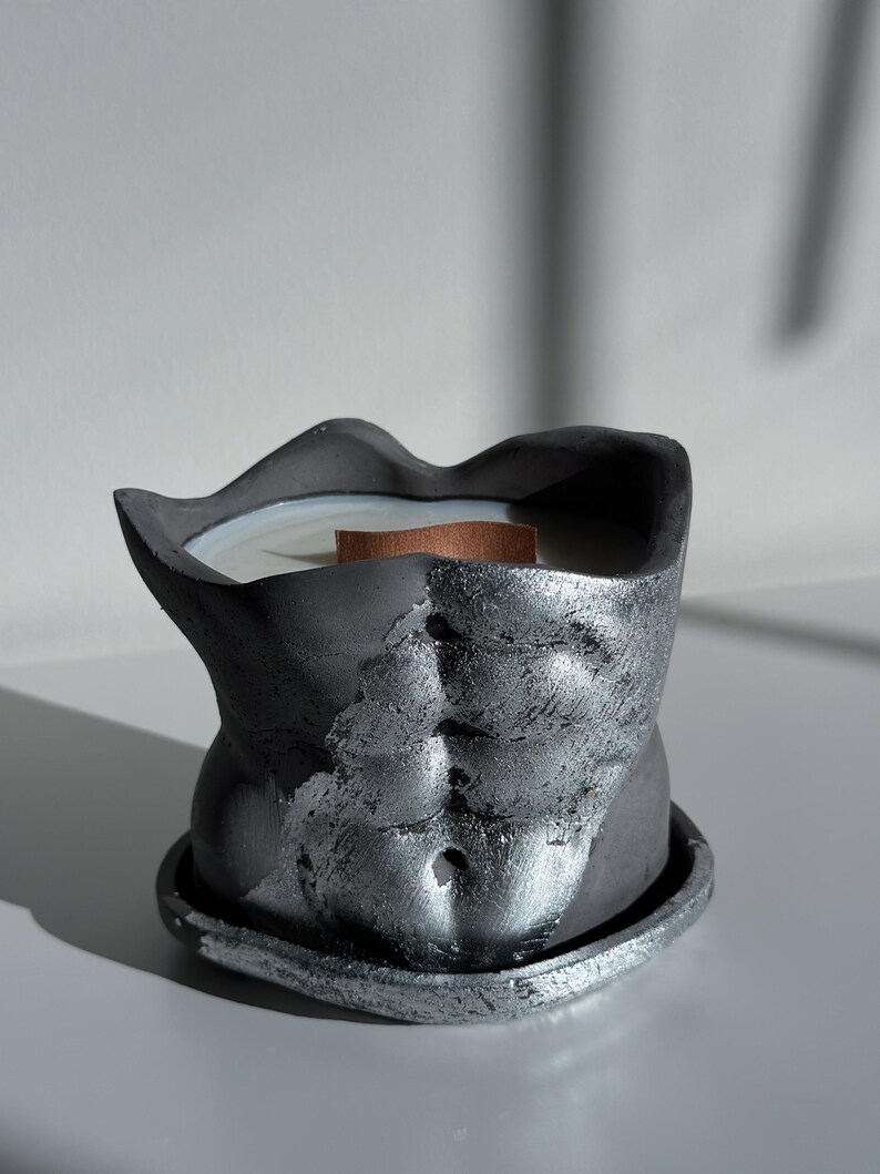 May include: A dark gray candle in the shape of a torso with a silver metallic finish. The candle has a wooden wick and sits on a matching base. The candle's design features sculpted details of the abdominal muscles.