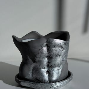 May include: A dark gray decorative planter shaped like a torso, with a textured, metallic silver finish on the front. The planter sits on a matching saucer. The interior appears to hold a white candle.