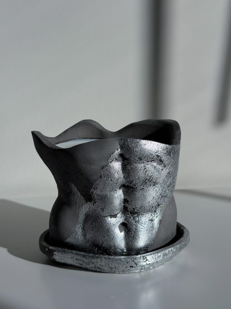 May include: A dark gray decorative planter shaped like a torso, with a textured, metallic silver finish on the front. The planter sits on a matching saucer. The interior appears to hold a white candle.