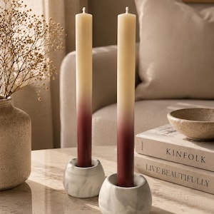 Peut inclure: Deux bougies ombrées dans des bougeoirs en marbre blanc. Les bougies passent du jaune pâle au bordeaux. Les bougeoirs ont une forme texturée et organique. Les bougies sont sur une table en marbre.