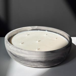 Puede incluir: Una vela grande y redonda en un cuenco de hormigón veteado gris y blanco. La vela tiene cera blanca cremosa con seis mechas. El cuenco se asienta sobre una superficie blanca con un fondo gris oscuro.
