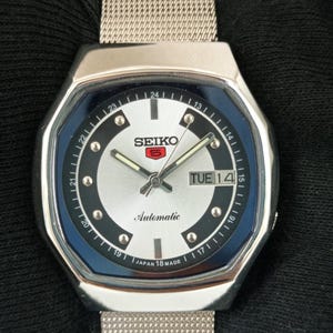 May include: A silver-toned Seiko 5 automatic wristwatch featuring a round silver dial with a dark blue outer ring, silver hour markers, luminous hands, and a day-date display at the three o'clock position. The watch has a stainless steel mesh band.