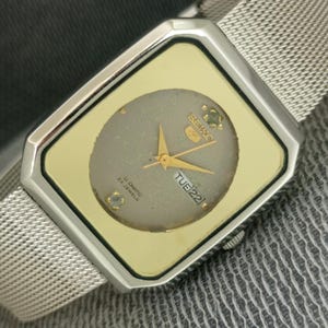 May include: A Seiko 5 automatic wristwatch with a rectangular silver-tone case and a gold-tone bezel. The dial features a circular grey center with gold hands, two small stone accents, and a day-date window displaying TUE 22. A silver mesh strap.