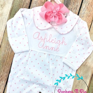May include: White baby onesie with pink polka dots and a pink bow. The onesie has a white collar with pink trim and the name "Ashleigh Anne" embroidered in pink.