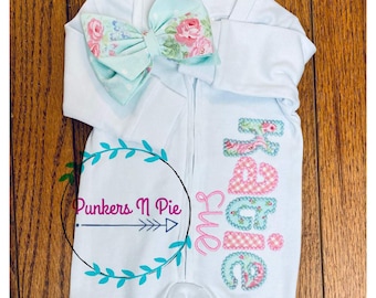 May include: A light blue baby onesie with the word "sweet" embroidered in pink and green floral fabric. The onesie has a zipper closure and foot coverings. A matching headband with a floral bow is included.