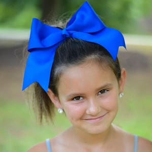 May include: A young girl with a bright blue bow in her hair smiles at the camera. She is wearing a gray tank top.